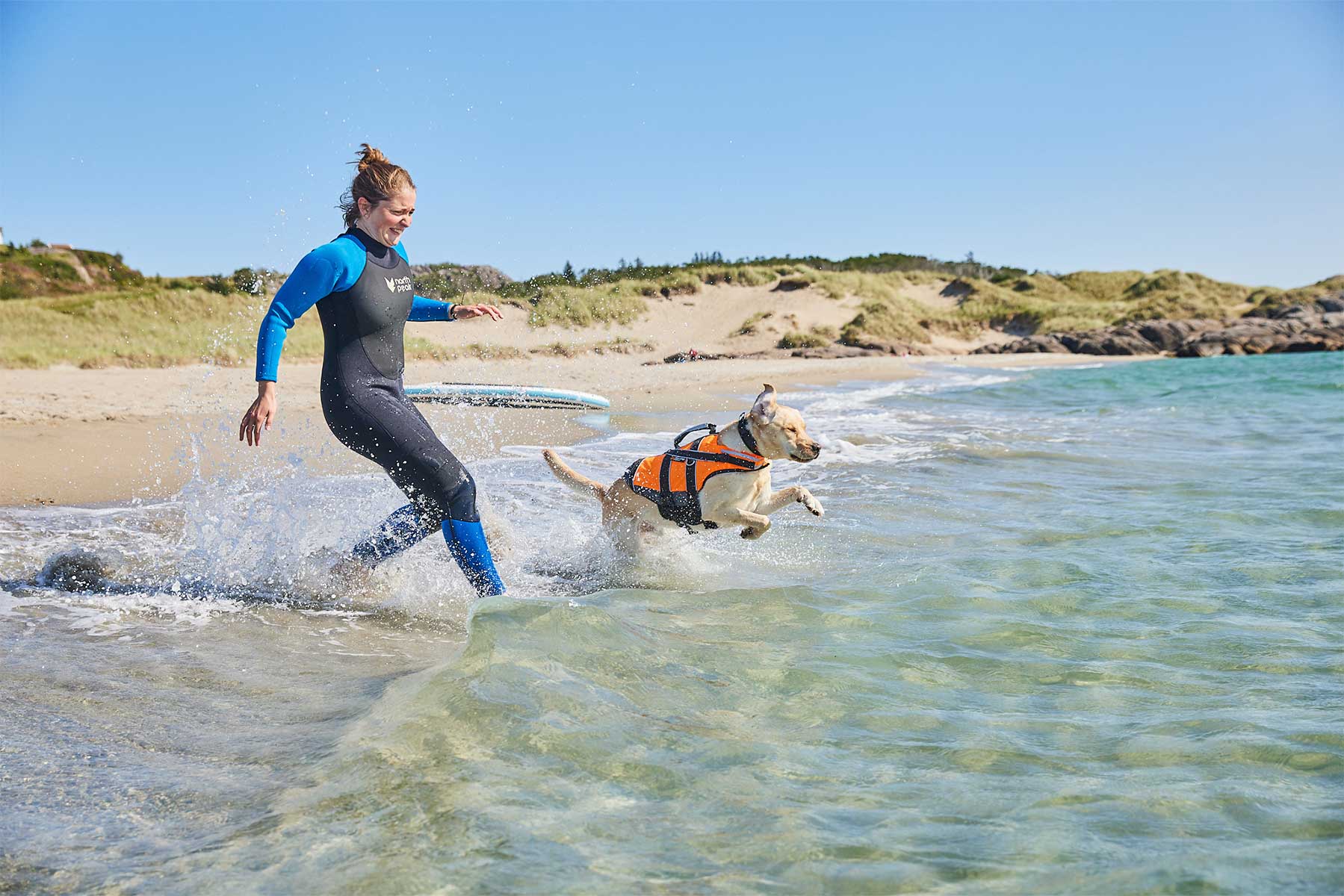 Chica con neopreno entrando a la playa con su perro con chaleco salvavidas naranja