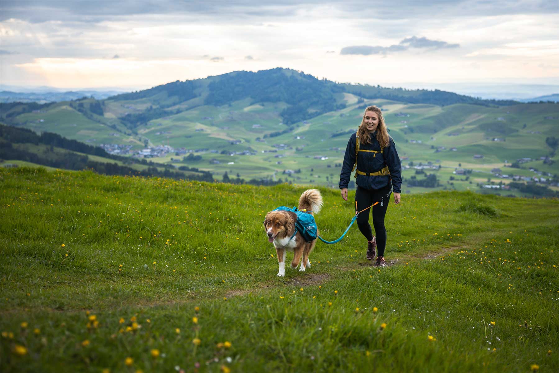 Chica paseando por una pradera verde con la mochila Non-stop dogwear Trail dog backpack y su perro