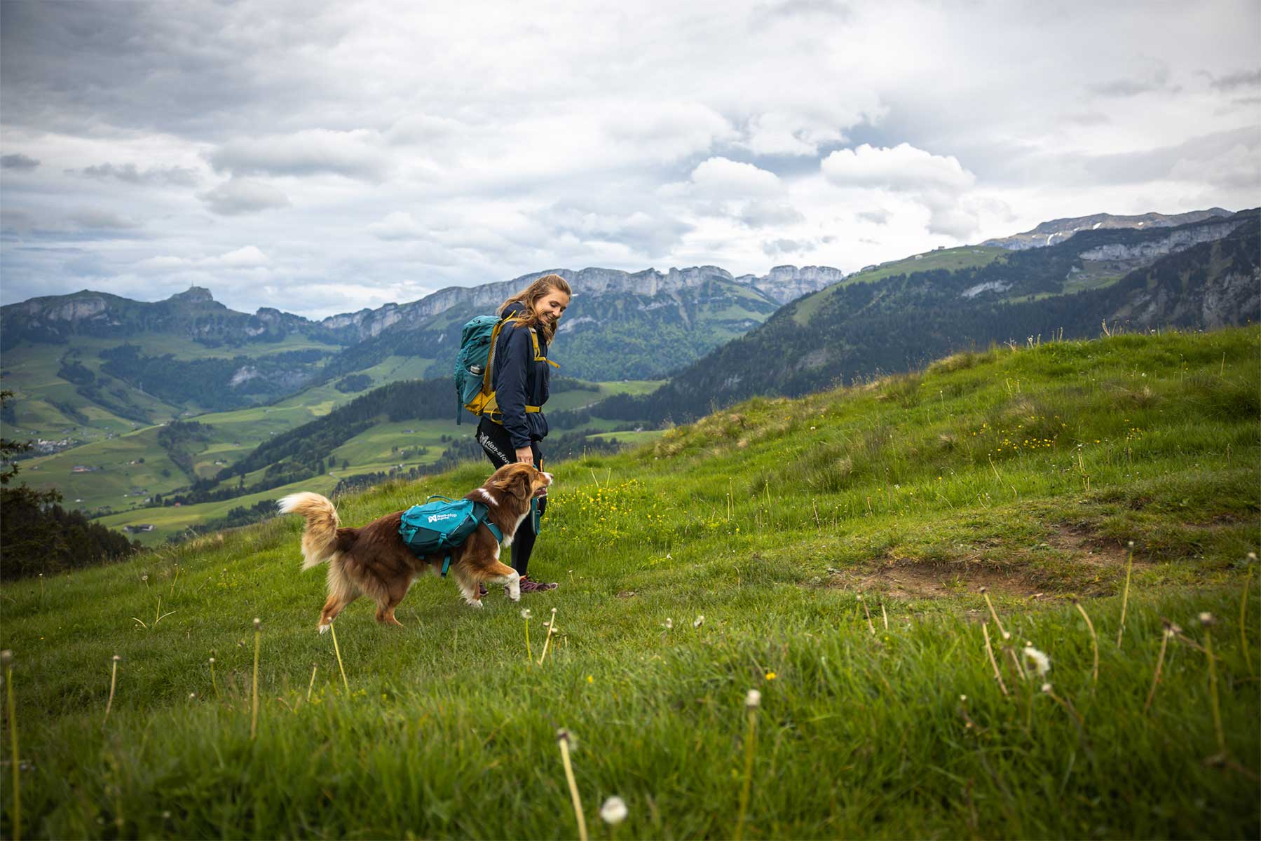 Chica paseando por una pradera verde con la mochila Non-stop dogwear Trail dog backpack y las montañas al fondo