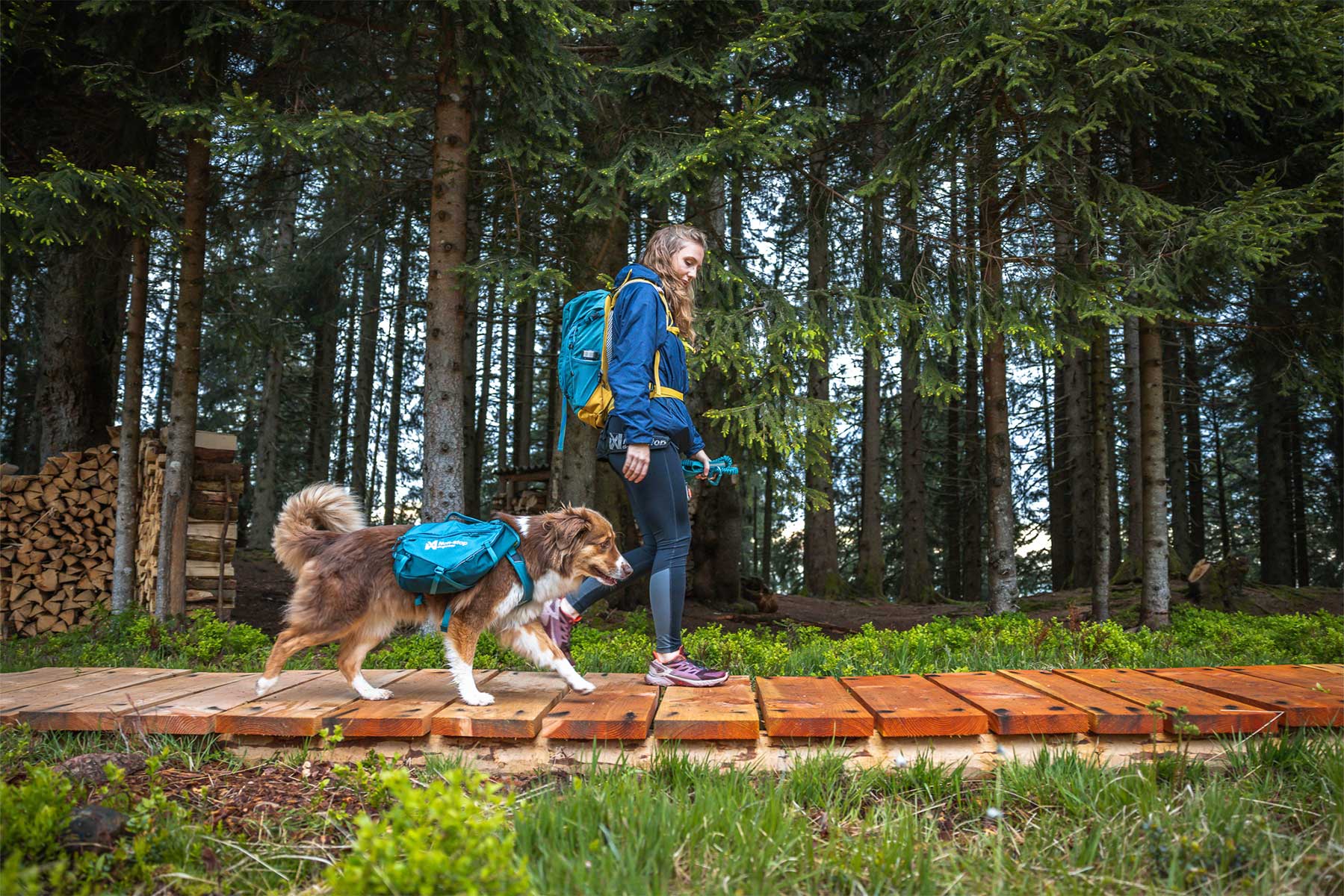 Chica atravesando un puente con la mochila Non-stop dogwear Trail dog backpack y su perro
